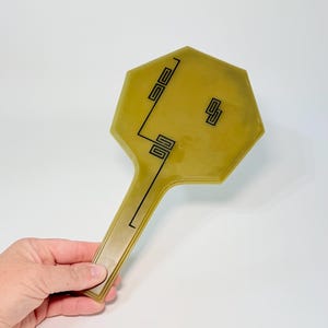 May include: An olive green hand mirror with an octagonal shape and a long handle. The surface features black geometric line patterns consisting of interconnected squares and rectangles arranged in a linear design across the face of the mirror.