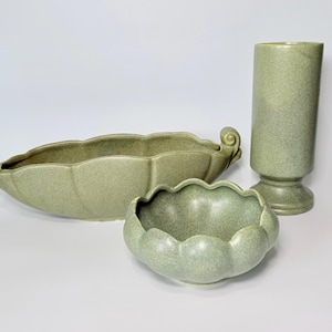 May include: A set of three ceramic decorative pieces in a speckled sage green color. The set includes a long, oval-shaped dish with scrolled handles, a small scalloped bowl, and a tall, cylindrical vase with a pedestal base.
