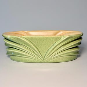 May include: A rectangular, Art Deco-style planter in a soft green hue. The planter features a fan-like design on the sides and a light beige interior. The overall shape is oval, and the planter is likely made of ceramic or a similar material.