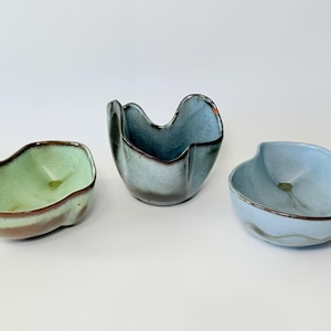 May include: Three ceramic bowls in shades of green and blue. The bowls have organic shapes with a dark brown rim. The centre bowl is a darker blue with a unique, folded design. The other two bowls are lighter shades of green and blue.
