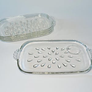 May include: Two clear glass serving trays with decorative handles. The trays are rectangular with rounded corners. One tray has a pattern of raised teardrop shapes. The other tray has a similar design.