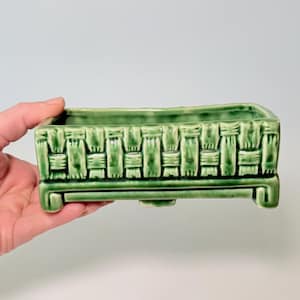 May include: A rectangular, green ceramic planter with a woven basket design. The planter is elevated on four small feet. The color is a vibrant, glossy green. This decorative planter is suitable for small plants or succulents.