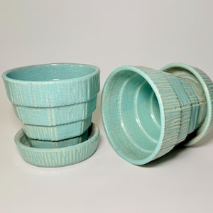 Peut inclure: Deux pots de fleurs en céramique turquoise avec soucoupes assorties. Les pots présentent un motif texturé à gradins avec des lignes verticales. Un pot est droit et l'autre est sur le côté. Les soucoupes ont un motif texturé similaire.
