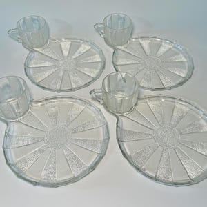 May include: Four clear glass teacup and saucer sets. Each set features a cup with a handle attached to a uniquely shaped saucer. The saucer has a sunburst design with textured detailing. The sets are arranged on a white surface.