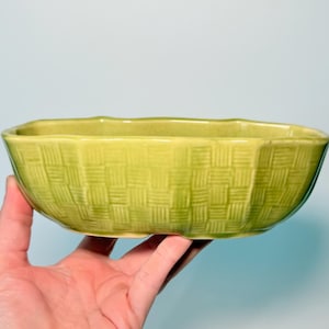 May include: An oval ceramic bowl in a light olive green color featuring a textured basket weave pattern on the exterior surface. The bowl has a smooth, glossy finish and a slightly scalloped rim, providing a decorative vessel for various uses.