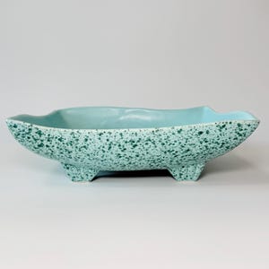 May include: A turquoise ceramic planter with a speckled pattern and three small feet. The oblong bowl has a wavy rim and a smooth interior. The exterior is covered in a speckled pattern of darker green.