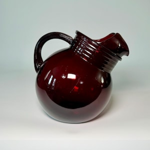 Peut inclure: Une carafe en verre rouge foncé avec un corps arrondi et une anse incurvée. La carafe a un bec verseur et un col côtelé. Le verre reflète la lumière, créant des reflets et des ombres. Cette carafe décorative est idéale pour servir des boissons.