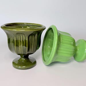 May include: Two green ceramic pedestal urns. One urn is upright, the other is on its side. Both urns have a ribbed design and a glossy finish. The urns are a shade of green, and are suitable for decorative purposes.