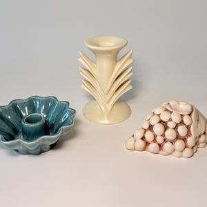 May include: Three ceramic candle holders in a studio setting. A teal scalloped candle holder, a cream-colored, fan-shaped candle holder, and a beige grape cluster candle holder are displayed on a white surface.