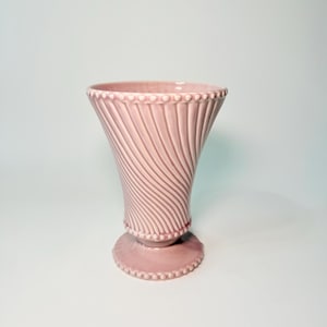 May include: A light pink ceramic vase featuring a flared opening and a pedestal base. The body of the vase is decorated with a series of diagonal, swirling ridges, while the rim and the base are accented with a delicate, raised beaded texture.