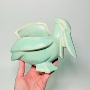 May include: A light green ceramic pelican-shaped wall pocket. The bird has a large beak and a pocket on its back. The ceramic piece is a light green colour with a glossy finish. The pelican is a decorative item.
