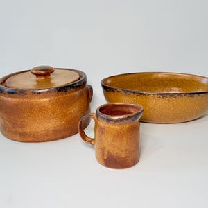 May include: A set of three brown ceramic kitchen items: a lidded casserole dish, a mug with a handle, and a large oval bowl. The items have a speckled texture and dark brown trim.