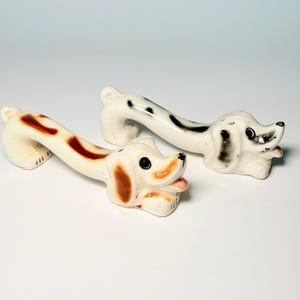 May include: Two ceramic dog-shaped shakers with elongated bodies. One shaker features brown spots and ears, while the other has black spots and ears. Both figures have black eyes, pink tongues, and small holes on their heads for dispensing contents.