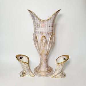 May include: A tall central vase and two smaller matching side vases, all featuring a white and gold distressed finish. Each piece has a flared, petal-like rim with gold trim and a textured, brush-stroke pattern across the exterior surface.
