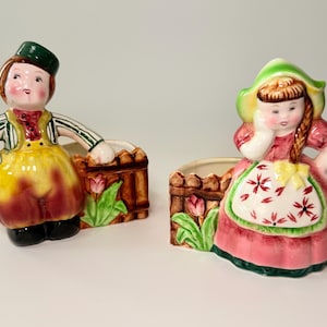 May include: A pair of vintage ceramic planters featuring figures in traditional attire. One figure wears a green hat and yellow overalls, the other a pink dress and apron. Each planter has a brown fence design with tulip accents.