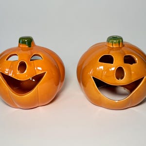 May include: Two orange ceramic jack-o'-lanterns with smiling faces. Each pumpkin has a green stem and cut-out eyes and mouth. The pumpkins are glossy and appear to be candle holders, perfect for Halloween decorations.
