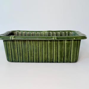 May include: A rectangular ceramic planter with a deep olive green glaze and a textured exterior that mimics the appearance of bamboo stalks. The vessel features a wide, flat rim and vertical ridges running along all four sides of the container.