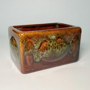 May include: A rectangular ceramic planter featuring a glossy brown glaze with a textured, mottled green drip pattern cascading from the top rim. The vessel has rounded corners and a smooth, reflective finish across its exterior surface.