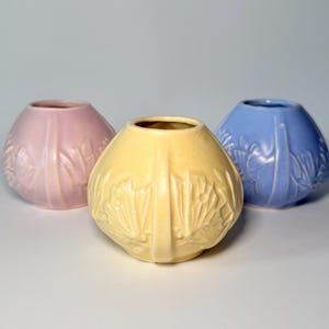 May include: Three ceramic vases in pastel shades: pink, yellow, and blue. Each vase has a rounded form with a textured, leaf-like design. The vases are displayed on a white surface.