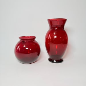 May include: Two ruby red glass vases. One is a round, bulbous shape, and the other is a taller, flared vase with a wider base. Both have a glossy finish and are set against a plain white background.
