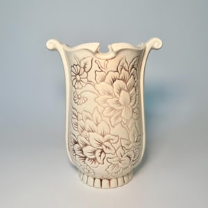 May include: Cream-colored ceramic vase with a floral relief pattern. The vase has a flared top with decorative handles and a pedestal base. The flowers are in a darker cream color, creating a contrast against the lighter background. The vase is approximately 10 inches tall.