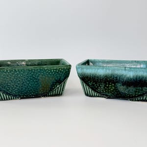 May include: Two rectangular ceramic planters. One is a deep teal green, the other a turquoise blue with dark green accents. Both have a textured surface and a curved, striped base. Suitable for succulents or small plants.