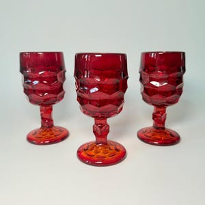 May include: Three ruby red vintage goblets with a geometric pattern on the bowl and a detailed stem. The base of the goblets is a gradient of orange to red. The glasses are set against a white background.