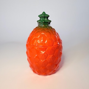 May include: A ceramic pineapple-shaped container with a lid. The container is a vibrant orange colour with a textured, geometric pattern. The top of the pineapple is green. The container is likely used for storage or decoration.