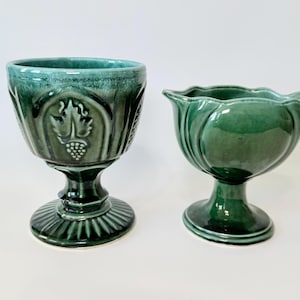May include: Two green ceramic goblets. One goblet has a detailed grape and leaf design. The other goblet has a tulip-shaped design. Both goblets have a pedestal base and are on a white background.
