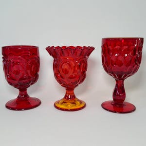 May include: Three ruby red glass goblets. The goblets have different designs, including embossed patterns and scalloped rims. The glass has a vintage aesthetic, with a rich, deep red color. The goblets are displayed on a white background.