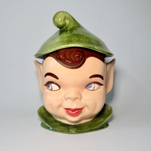 May include: A vintage ceramic cookie jar shaped like a smiling elf's head. The jar features a green pointed hat, brown hair, and a rosy complexion with blue eyes and red lips. The elf has large ears and a green collar.
