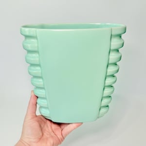 May include: A mint green ceramic planter with a rectangular shape and a ribbed design on the sides. The planter is held by a hand against a white background. The smooth, glossy finish gives it a vintage aesthetic.