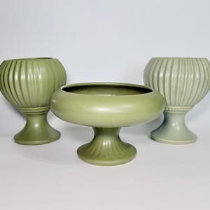May include: Three green ceramic vases of varying shapes and sizes. Two vases have a ribbed design and pedestal base. The third vase is a shallow bowl shape with a pedestal base. All three vases are in shades of green.