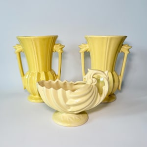 May include: Three vintage yellow ceramic vases. Two tall vases with handles and leaf-like accents, and a smaller, shell-shaped bowl. The vases have a ribbed design and are set against a light background. A decorative set for home decor.