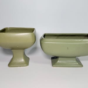 May include: Two sage green ceramic planters. The planters have a square opening and a pedestal base. The planter on the left is taller than the one on the right. The planters are empty and ready to be filled with plants.