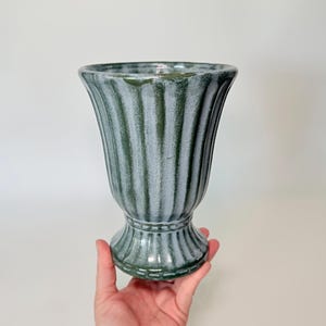 May include: A green and white ceramic vase with a ribbed design. The vase has a pedestal base and a flared top. The vase is held by a hand against a white background. The vase is approximately 8 inches tall.