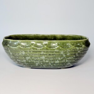May include: An oval ceramic bowl featuring a deep moss green glaze with a speckled, lighter green finish. The exterior surface is decorated with a horizontal ribbed texture and a scalloped pattern along the upper rim, creating a distinct relief.