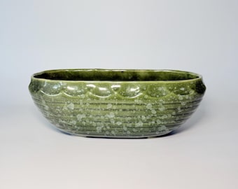 May include: An oval ceramic bowl featuring a deep moss green glaze with a speckled, lighter green finish. The exterior surface is decorated with a horizontal ribbed texture and a scalloped pattern along the upper rim, creating a distinct relief.