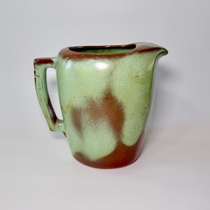 May include: A vintage ceramic pitcher with a unique color scheme. The pitcher features a light green base with brown accents and a brown rim. It has a handle and a spout for easy pouring. This decorative pitcher is a great addition to any kitchen.
