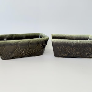 May include: Two rectangular ceramic planters. One is decorated with a grape design, the other with a floral pattern. Both have a dark brown base with a green and brown rim.