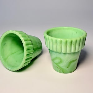 May include: Two small, light green glass flower pots. One pot is upright, the other is on its side. Both have a ribbed rim and a swirled pattern. These decorative pots are ideal for small plants.