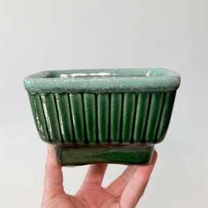 May include: A rectangular, green ceramic planter with vertical ribbed detailing. The planter has a square base and a slightly textured, light green rim. The object is held in a hand against a plain white background.