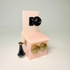 May include: A vintage-style pink telephone-shaped salt and pepper shaker set. The set features a black receiver, gold-colored dials, and a black earpiece. The shaker set is a decorative kitchen item.