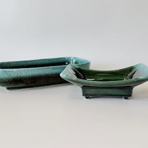 May include: Two ceramic serving dishes. One is a long, rectangular shape, and the other is a square shape with curved sides. Both are glazed in shades of teal and green, with a glossy finish. The dishes are empty and sit on a white surface.