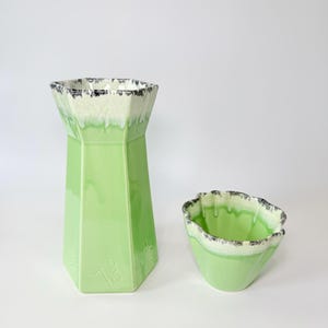 May include: Two-piece ceramic set in a light green hue. The taller piece is a vase with a geometric design, and the smaller piece is a bowl. Both have a white and black trim around the top edge. The vase has the number "13" on the side.