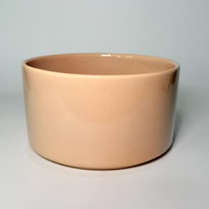 May include: A cylindrical ceramic bowl with a smooth, matte finish in a solid light peach or soft beige color. The bowl features straight, vertical sides and a flat base, presenting a clean, uniform appearance with no additional patterns or textures.