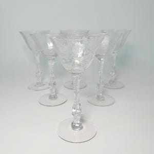 May include: A set of six clear, etched glass cocktail glasses. Each glass has a wide bowl, a slender stem with decorative detailing, and a circular base. The glasses are arranged in a semi-circular pattern against a white backdrop.