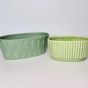 May include: Two ceramic planters in shades of green. The larger, oval-shaped planter has a repeating oval pattern. The smaller, rectangular planter has vertical ribbed detailing. Both are on a white background.