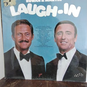 May include: A vintage vinyl record album titled "Rowan & Martin's Laugh-In." The album cover features two men in tuxedos against a light blue background. The album's tracklist is visible on the left side of the cover.
