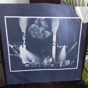 May include: A black and white concert poster featuring a band performing on stage. The poster has a dark blue background with a white border. The band members' faces are visible at the top, and the stage is lit with spotlights.